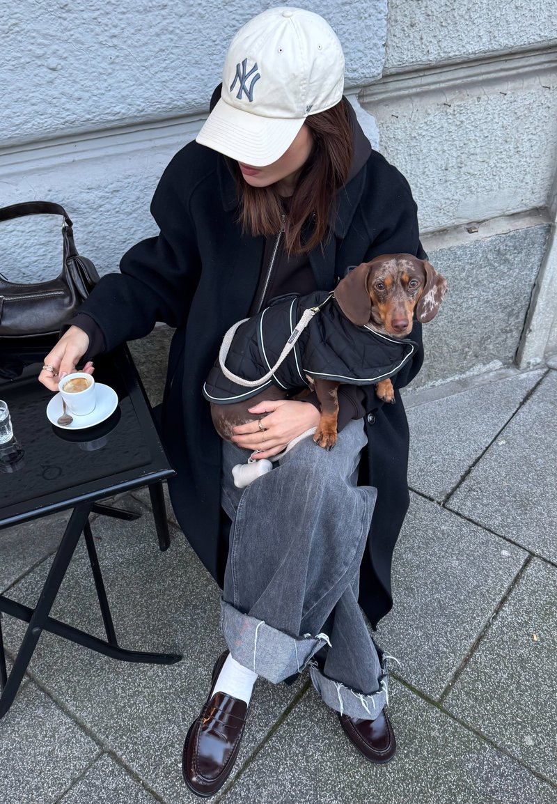 Black coat over a dark jumper, grey jeans with frayed hem, brown loafers, and a beige cap. Holding a brown dog in a black coat, sipping coffee.