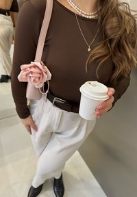 Maglietta marrone a maniche lunghe, pantaloni bianchi a vita alta, borsa rosa con dettaglio a rosa, tiene una tazza bianca e indossa una collana di perle.