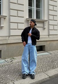 Fekete, gombos ing fehér póló fölött, laza világoskék farmernadrággal és masszív fekete cipőkkel párosítva. Városi kültéri környezet.