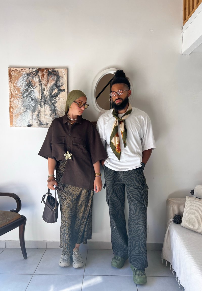 Due persone posano all'interno. La donna indossa una camicia oversize marrone scuro, una gonna stampata e un hijab verde oliva. L'uomo indossa una camicia bianca, pantaloni con fantasia e sneakers verdi.