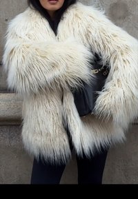 Personne portant un manteau en fourrure blanche duveteuse et un pantalon noir, tenant un sac à main noir devant un mur en pierre.