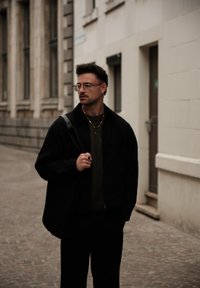 Zwarte jacket over een donkergroen trui, zwarte broek en een bril. Opvallende accessoires zijn gouden kettingen en een ring aan de hand.