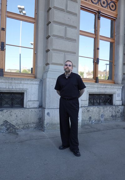 Hombre vestido con ropa negra y gafas de sol, de pie con las manos detrás de la espalda junto a un edificio de piedra con ventanas grandes que reflejan el cielo.