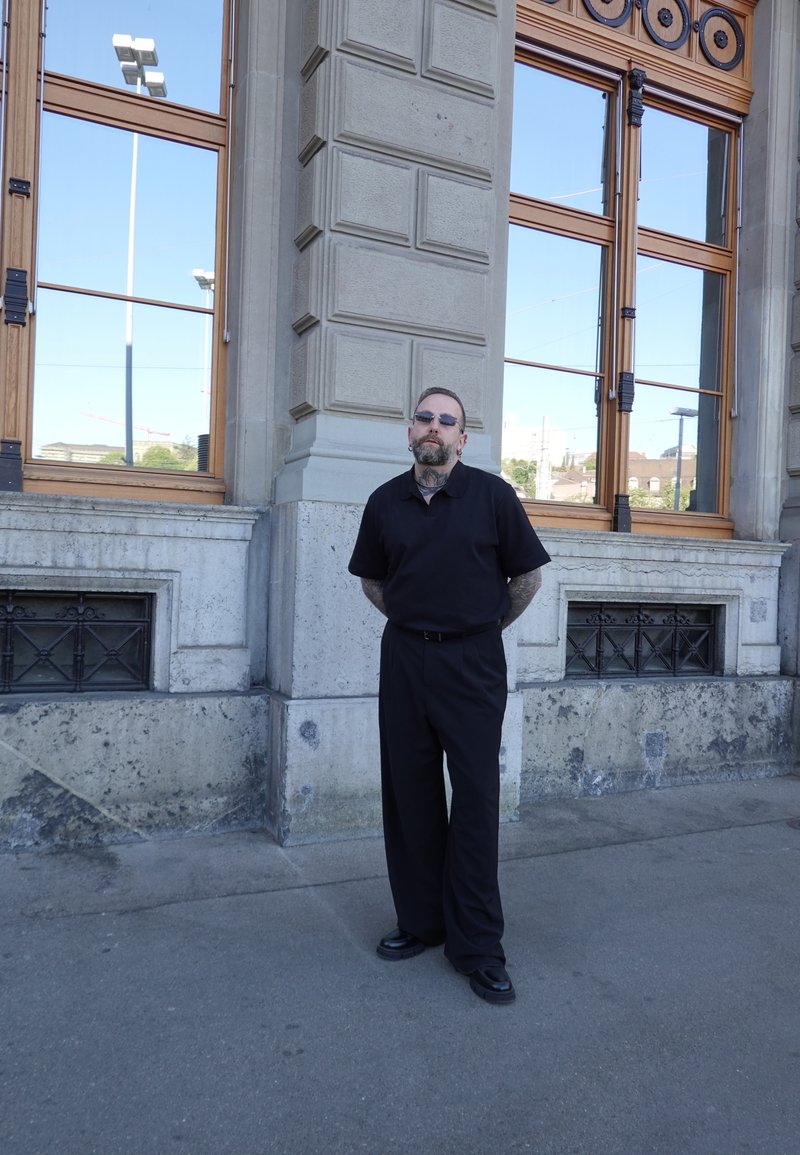 Mann in schwarzer Kleidung und Sonnenbrille, steht mit den Händen hinter dem Rücken vor einem Steingebäude mit großen Fenstern, die den Himmel spiegeln.