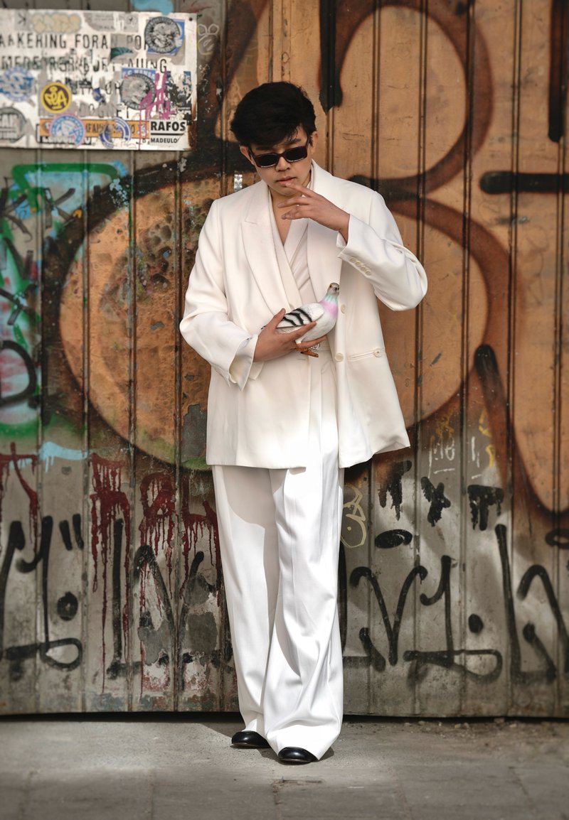 Homme en costume blanc et lunettes de soleil tenant un pigeon devant un mur en bois couvert de graffitis.