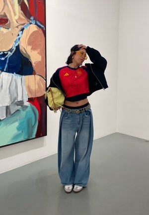 Persona con vaqueros anchos, camiseta deportiva corta roja, gafas de sol amarillas, sosteniendo una bolsa, posando con la mano en la cabeza junto a una pintura grande y colorida en una galería.