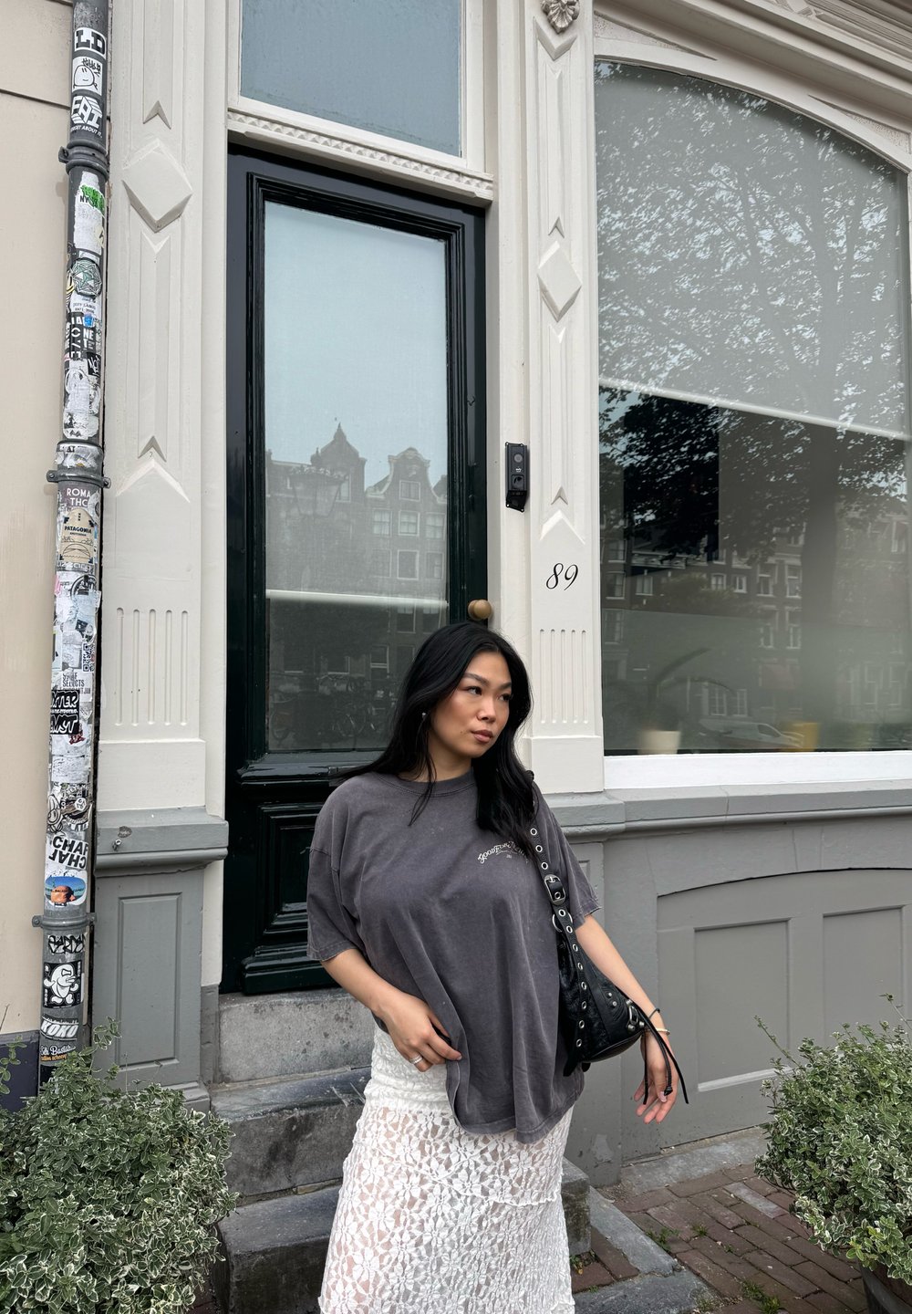 Overmaat grijze t-shirt gecombineerd met een witte kanten rok. Zwarte handtas met zilveren hardware. Groene bladeren en een decoratieve deur zijn zichtbaar.