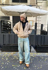 Beige zip-up hoodie over een zwarte hoodie, lichtblauwe wijde jeans en tan laarzen; staande onder een grote crèmekleurige paraplu met een houten achtergrond.