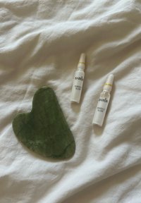Strumento gua sha in giada verde a forma di cuore, con delle marcature; due fiale di vetro bianche etichettate "BABOR PERFECT GLOW" con accenti gialli accanto.