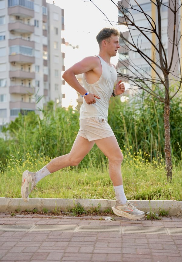 Joven corriendo al aire libre por un camino pavimentado cerca de césped y edificios, vistiendo ropa deportiva ligera y zapatillas.