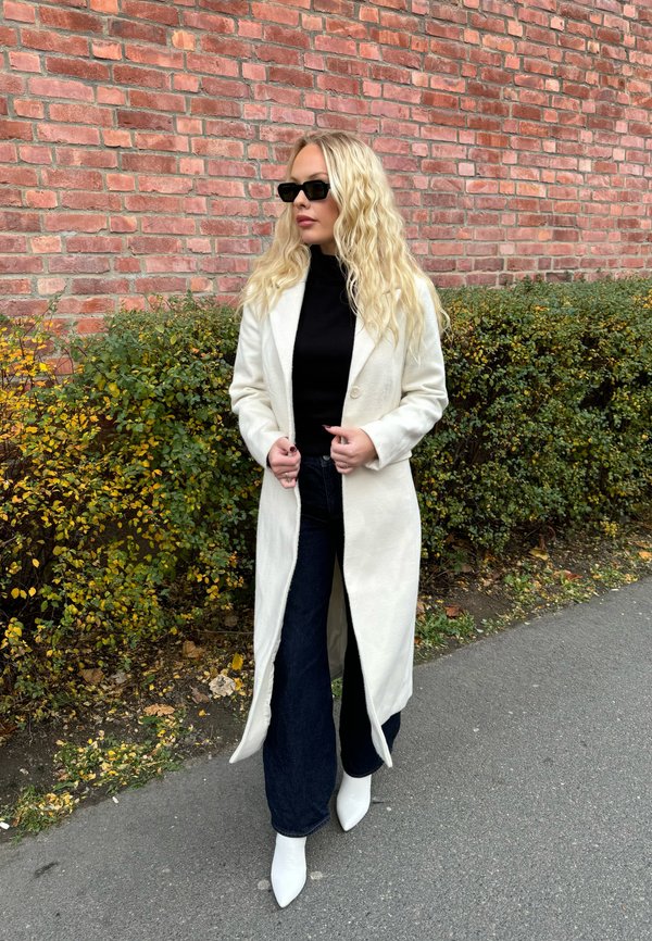 Long manteau blanc avec une finition texturée, col roulé noir, jeans évasés en denim et bottes blanches à bout pointu ; le tout devant un mur de briques.