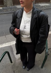 Personne aux cheveux courts portant une veste en cuir noire, un t-shirt blanc avec un motif graphique, un pantalon noir, des bagues en argent, et tenant un sac noir tressé, debout sur le trottoir.