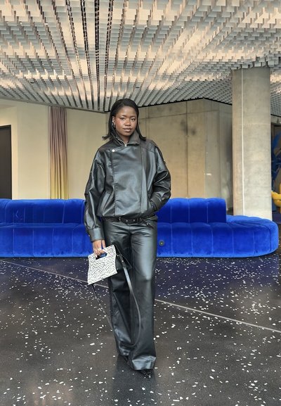 Mujer con chaqueta y pantalones de cuero negros, sosteniendo un pequeño bolso con estampado, de pie en una habitación moderna con sofá de terciopelo azul y techo texturizado.