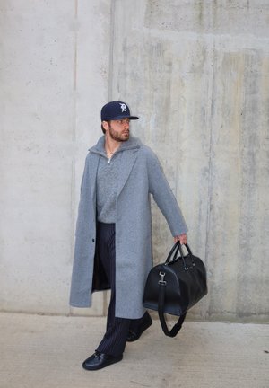 Hombre con abrigo gris, suéter, pantalones de rayas y gorra azul marino camina sosteniendo una bolsa de viaje negra frente a una pared de concreto.