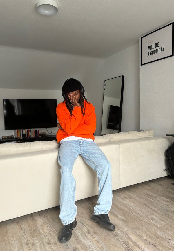 Uma pessoa sentada veste um hoodie laranja, uma t-shirt branca, jeans azul claro e botas pretas. O ambiente tem um sofá de cor clara e um espelho.