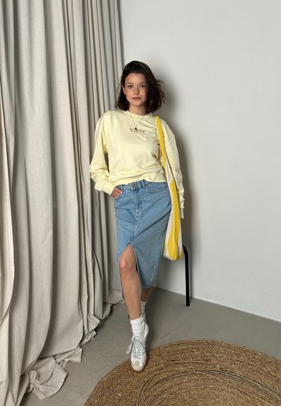 Camisa amarilla de manga larga con logo, falda de mezclilla azul clara con una abertura lateral, zapatillas blancas y un bolso amarillo y blanco. Fondo natural.