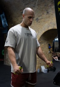 Mann i en lys grå t-skjorte med grafisk trykk, holder grønne håndtak på treningsutstyr, iført røde shorts med hvite striper; treningsstudio.