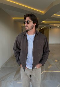 Giubbotto marrone con zip e tasca, indossato su una maglietta bianca. Pantaloni beige chiaro e occhiali da sole completano l'outfit, in un ambiente interno.
