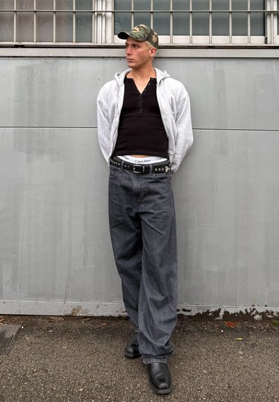 Sudadera gris con capucha sobre un henley negro, jeans gris holgados con un cinturón negro y zapatos negros, complementados con una gorra de camuflaje.