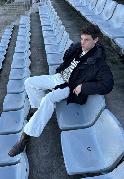 Joven con chaqueta negra, camiseta de rayas, pantalones blancos y botas recostado sobre asientos grises vacíos de un estadio.