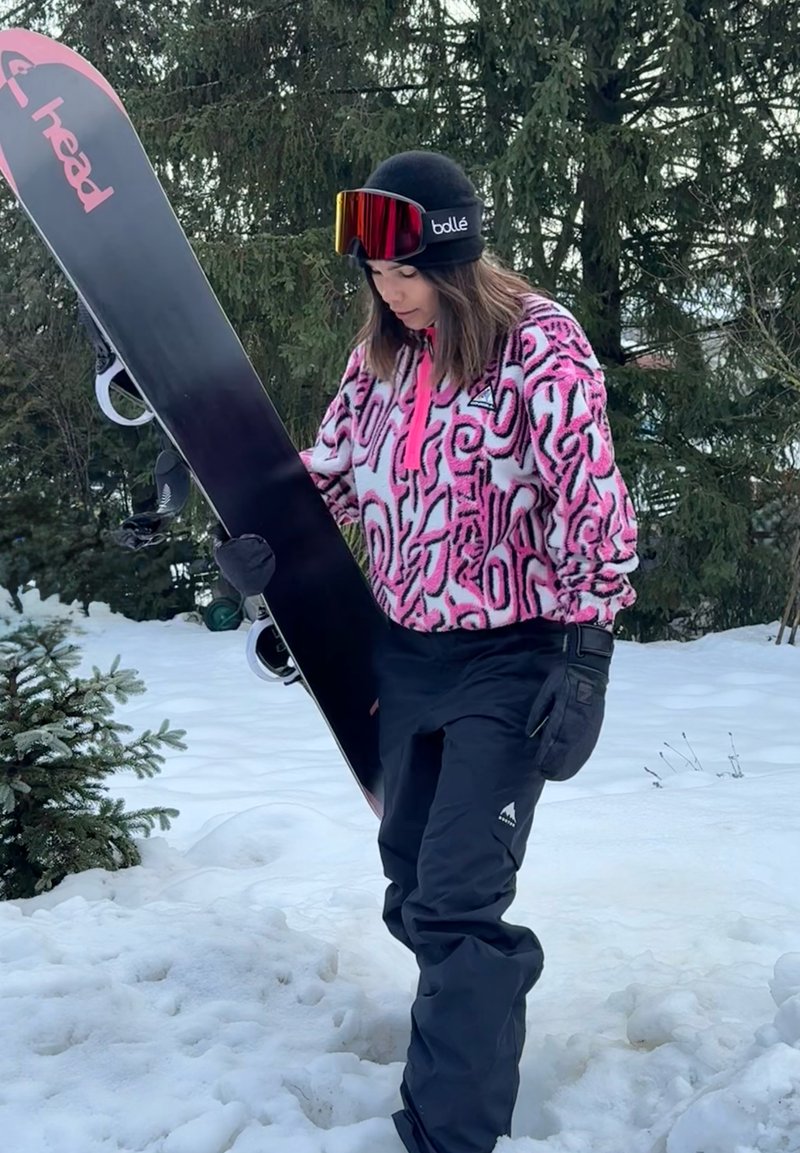 Person wearing a pink patterned jacket and black snow trousers holding a black snowboard in a snowy forest setting.