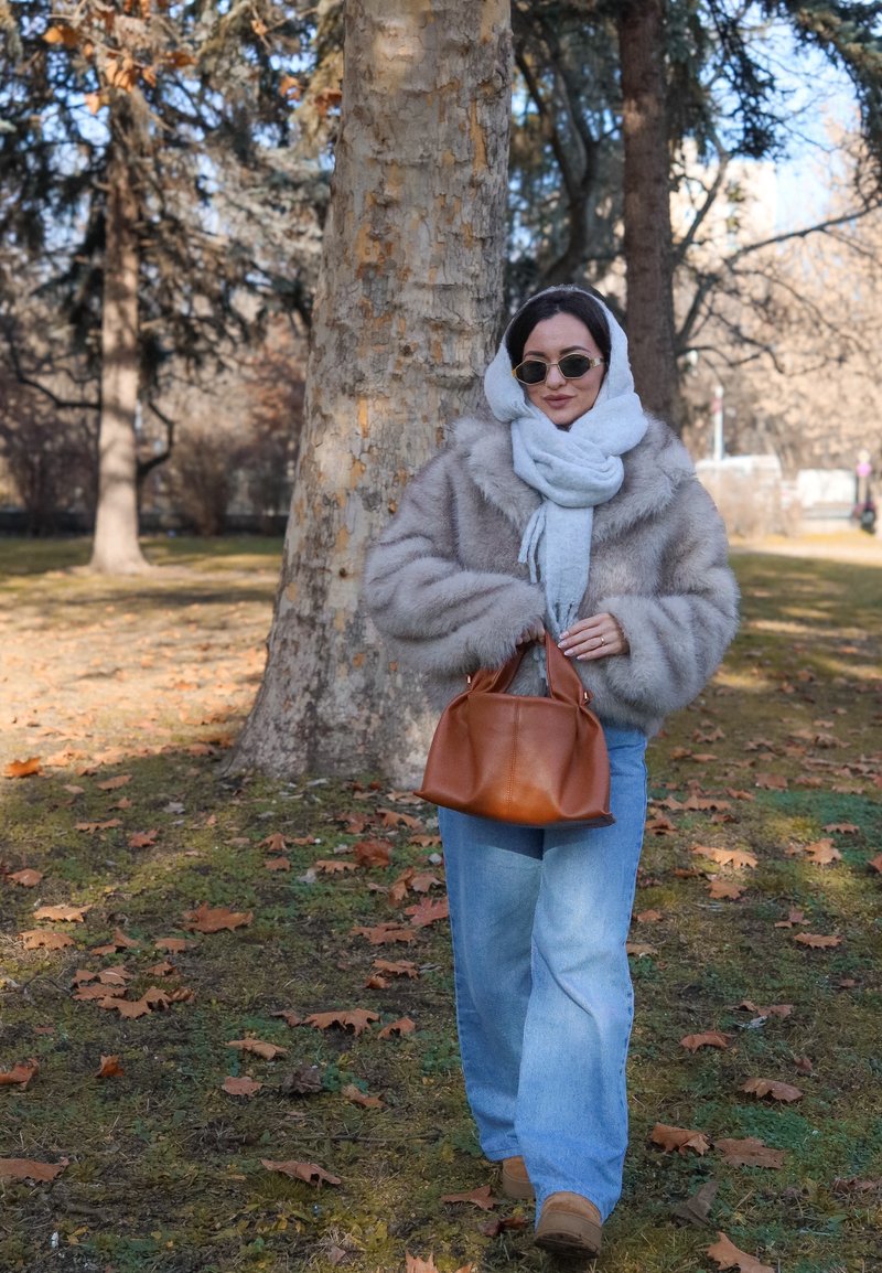 Giacca in pelliccia sintetica beige, sciarpa grigia, occhiali da sole rotondi, jeans a gamba larga azzurri, borsa marrone, che cammina su un'area erbosa con foglie cadute.