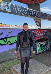 Čierna kožená bomber bunda a sukňa, priliehavý rolák, čierne pančuchy a členkové topánky, stojaca pred stenou pokrytou graffiti.