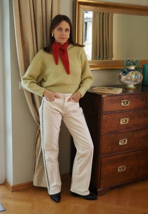 Knit green jumper, red scarf, cream wide-leg trousers with black side stripes, black shoes, standing next to wooden furniture and decor.