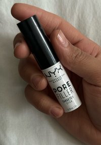 Käsi, joka pitää NYX Professional Makeup Pore Filler Targeted Stickiä, mustalla kannella ja valkoisella etiketillä kevyellä kankaan taustalla.