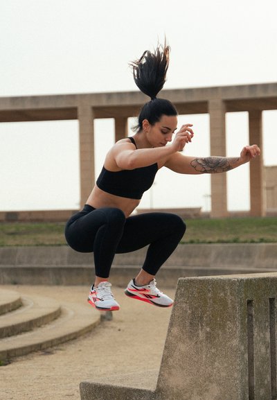 Atleta con top corto negro y leggings salta sobre una barrera de piedra gris, vistiendo zapatillas deportivas blancas, rojas y negras, con un tatuaje visible en el brazo.