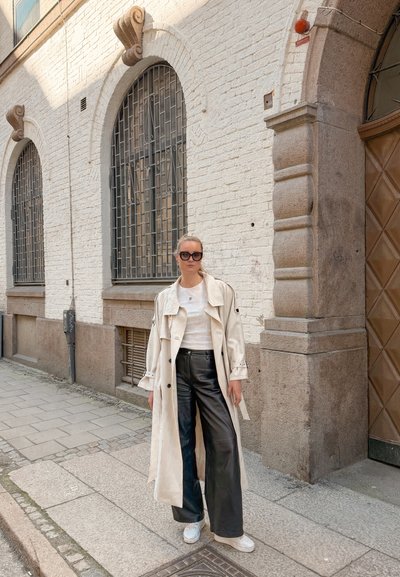 Mujer con un abrigo largo beige tipo trench, pantalones negros de pierna ancha, zapatillas blancas y gafas de sol, de pie en una acera empedrada junto a ventanas arqueadas.