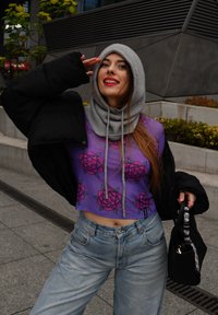 Μωβ κοντό τοπ με ροζ λουλουδάτο μοτίβο, το οποίο έχει επιπλέον μια μαύρη γεμιστή ζακέτα και γκρι πλεκτή κουκούλα, σε συνδυασμό με ψηλόμεσα ανοιχτόχρωμα τζιν.