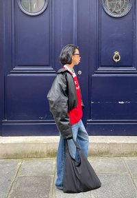 Zwarte leren oversized jack, rode shirt, blauwe jeans en een zwarte tote bag. Achtergrond met een donkerblauwe deur met ronde ramen.