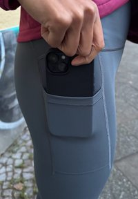 Szürke sport leggings oldalzsebbel, amelyben egy fekete okostelefon van. Az anyag rugalmasnak tűnik, jól látható varrási részletekkel.
