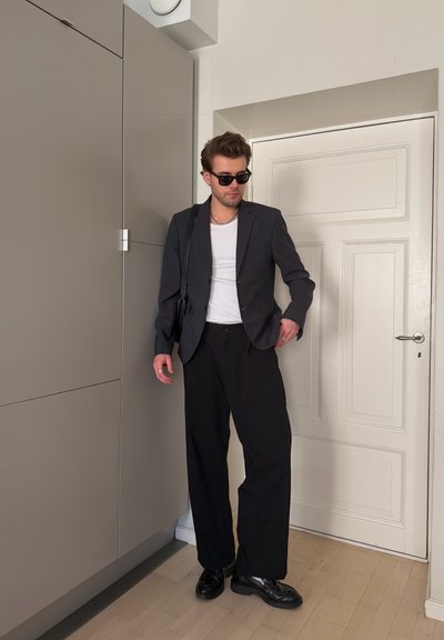 Hombre con gafas de sol negras, blazer oscuro, camiseta blanca, pantalones negros de pierna ancha y zapatos negros, de pie en el interior junto a una puerta blanca.