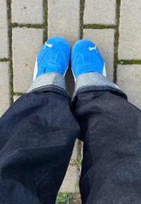 Blå ruskindssneakers med hvide detaljer, kombineret med mørke denimbukser. Synlig opsmøg ved anklen. Underlaget er en belagt stensti.