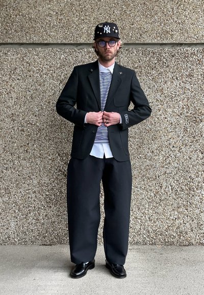 Blazer negro con un solo botón, camisa a rayas debajo, pantalones negros oversized y zapatos negros brillantes; acentuado con una gorra con tachuelas.