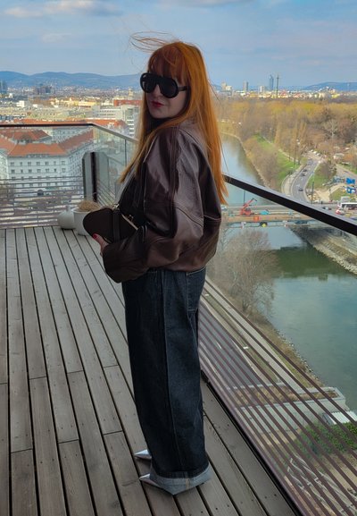 Persona pelirroja con gafas de sol, chaqueta de cuero marrón y vaqueros anchos, de pie en un balcón de madera con vistas a un río y paisaje urbano.