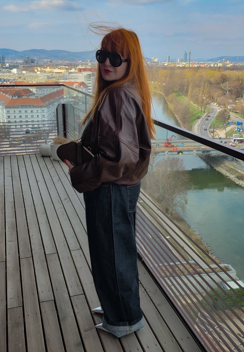 Άτομο με κόκκινα μαλλιά, γυαλιά ηλίου, καφέ δερμάτινο μπουφάν και φαρδιά τζιν στέκεται σε ξύλινο μπαλκόνι με θέα ποτάμι και αστικό τοπίο.