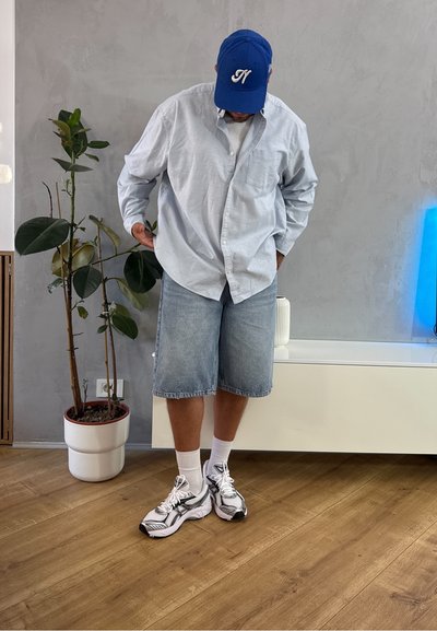 Camisa de botones azul claro, pantalones cortos de mezclilla claros, calcetines blancos y zapatillas negras y blancas. Planta en maceta y decoración moderna de fondo.