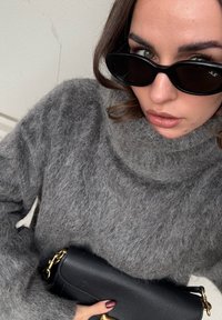 Szürke bolyhos pulóver magas gallérral. Fekete napszemüveg. Egy kis fekete textúrált kézitáskát tart, arany lánccal díszítve.