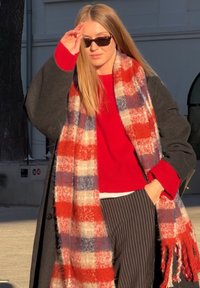 Femme portant des lunettes de soleil, un pull rouge, un long manteau gris et une grande écharpe à carreaux rouge, blanc et bleu, debout à l'extérieur au soleil.