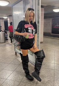 Mustikas mustas graafilises T-särgis ja mini-seelikus kannab kuni põlveni ulatuvaid musti kummisaapaid. Neil on rohelise joogi ja must koti.