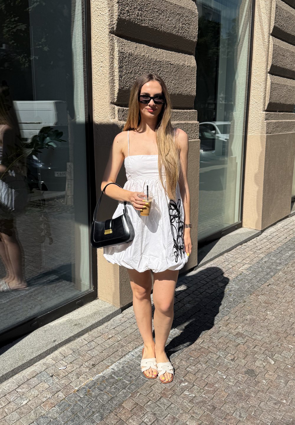 Hvid kjole med en stropløs, tætsiddende bodice og løs nederdel, detaljeret med sort broderi; parret med en sort håndtaske og lyse sandaler.