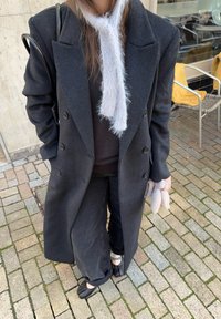 Lang sort uldfrakke med dobbeltradede knapper, båret over en sort top og sorte bukser med vide ben. Grå strikket halstørklæde, sorte sko.