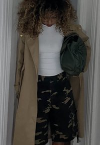 Jasnobeżowy trencz na białym dopasowanym topie, w połączeniu z czarnymi szortami w kamuflażu. Osoba trzyma zieloną torbę, stoi w pomieszczeniu.