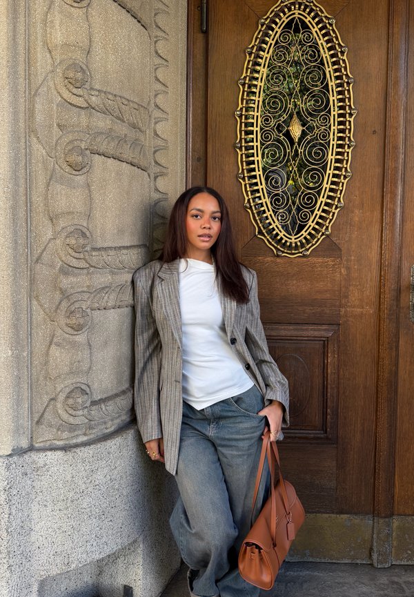 Mujer con blazer gris y vaqueros se apoya en una pared de piedra ornamentada junto a una puerta de madera marrón con una rejilla metálica oval decorativa.
