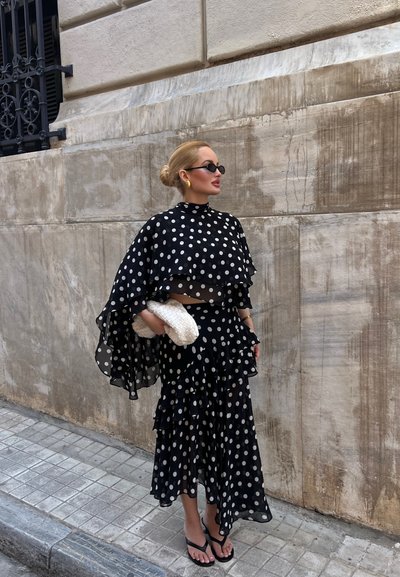 Mujer con capa y falda de lunares en blanco y negro, sosteniendo un bolso blanco tejido, de pie en la acera de la ciudad contra una pared de piedra.
