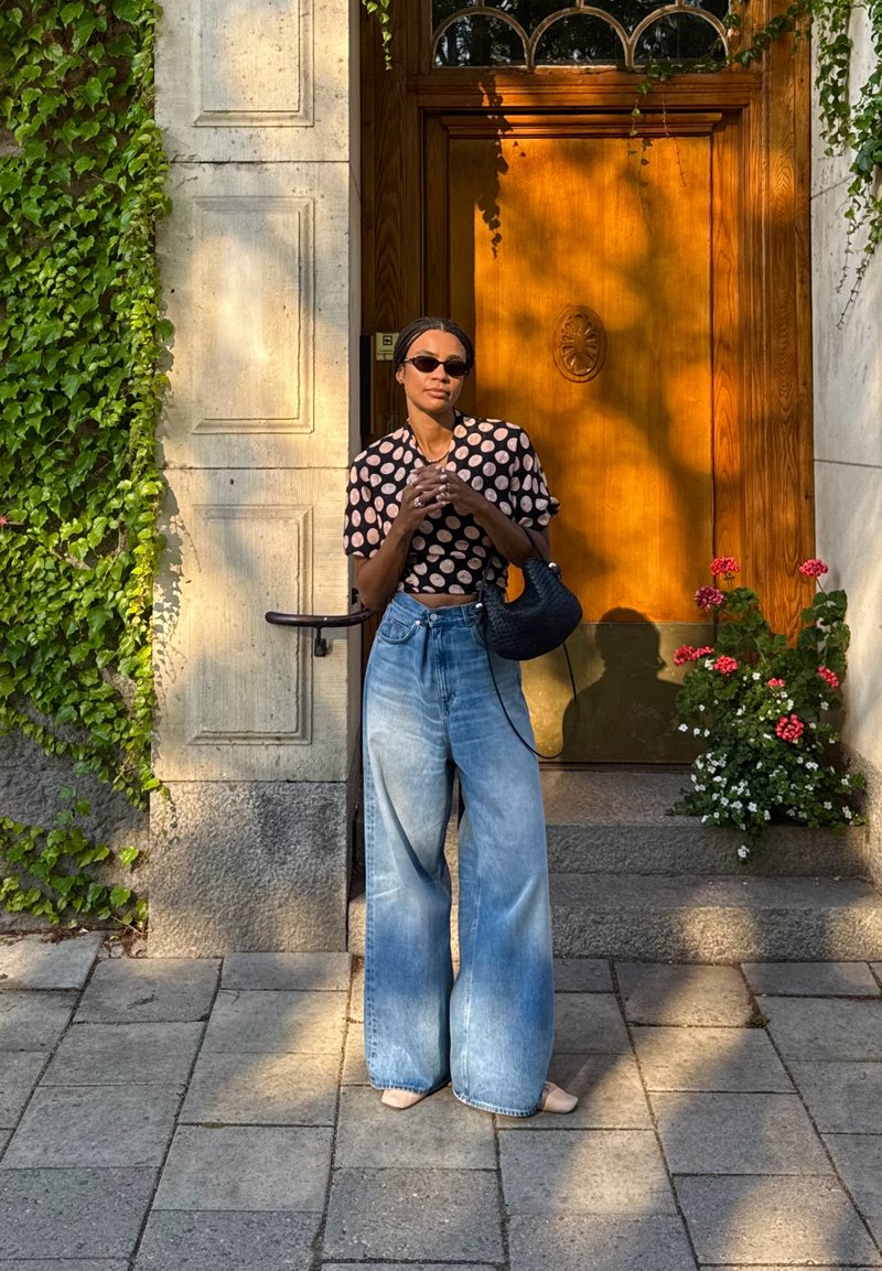 Donna con occhiali da sole, top a pois e jeans a gamba larga in piedi vicino a una porta di legno con edera e vasi di fiori alla luce del sole.