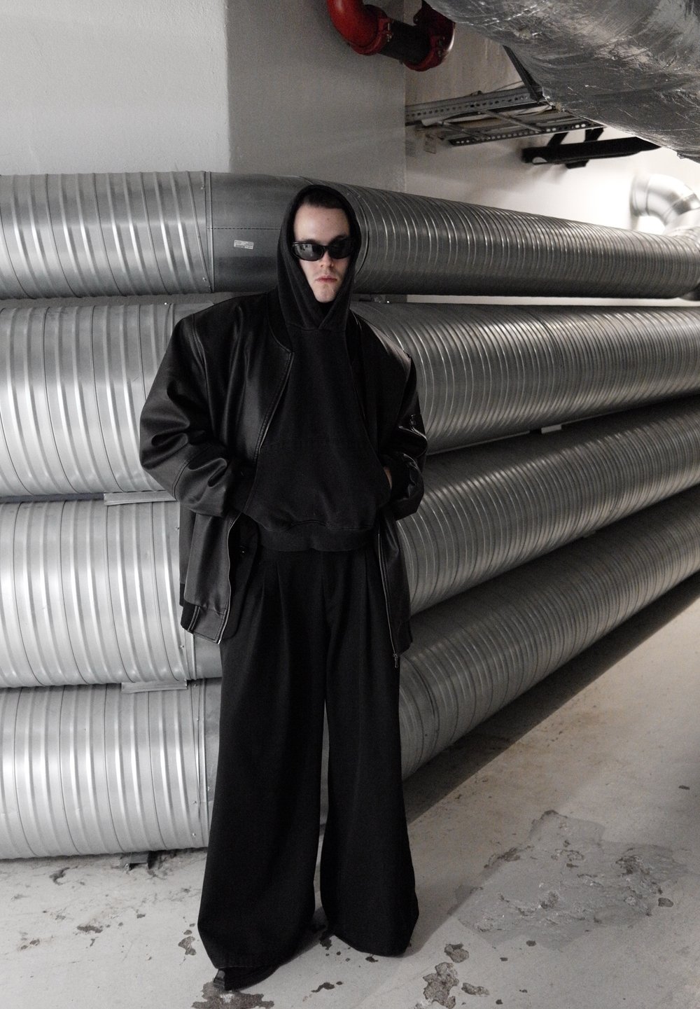 Svart oversize läderjacka, svart hoodie och vida svarta byxor. Modellen står mot silverfärgade ventilationsrör i en minimalistisk miljö.
